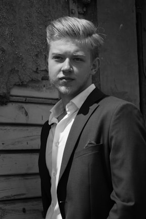 A man in a suit stands confidently in front of a textured, aged wooden background. The light highlights his facial features and well-groomed hair, giving a classic and timeless feel.