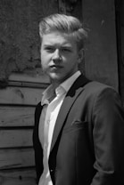 A man in a suit stands confidently in front of a textured, aged wooden background. The light highlights his facial features and well-groomed hair, giving a classic and timeless feel.