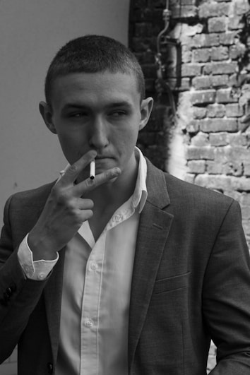 A person wearing a suit jacket and an unbuttoned dress shirt is holding a cigarette close to their lips. The background includes a brick wall with visible wiring.