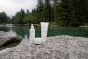 Skincare products are placed on a large rock near a serene lake surrounded by a dense forest. The water is clear, reflecting the green hues of the surrounding trees, creating a tranquil and natural setting.