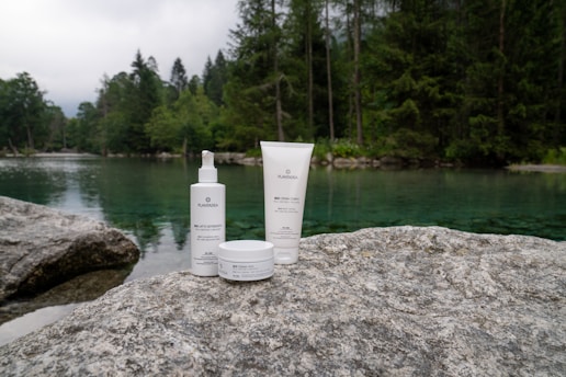 Skincare products are placed on a large rock near a serene lake surrounded by a dense forest. The water is clear, reflecting the green hues of the surrounding trees, creating a tranquil and natural setting.