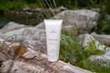 A cosmetic tube labeled 'Plantae Bio Crema Corpo' is placed on a log surrounded by natural vegetation and rocks, depicting an outdoor setting.