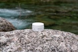 A jar of face cream labeled 'Bio Crema Viso' by PLANTADEA is placed on a large, textured rock. The background features a flowing stream with greenish water and blurred natural surroundings.