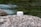 A jar of face cream labeled 'Bio Crema Viso' by PLANTADEA is placed on a large, textured rock. The background features a flowing stream with greenish water and blurred natural surroundings.
