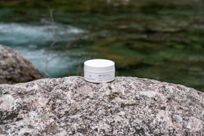 A jar of face cream labeled 'Bio Crema Viso' by PLANTADEA is placed on a large, textured rock. The background features a flowing stream with greenish water and blurred natural surroundings.