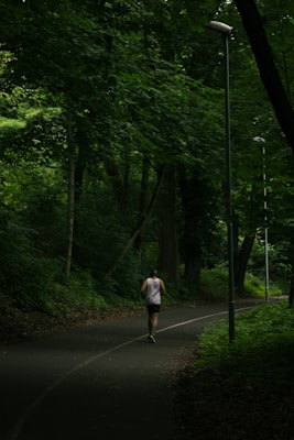 A person is jogging along a winding path surrounded by dense, lush green trees. The path is lined with street lamps and the area appears quiet and secluded, creating a serene atmosphere.