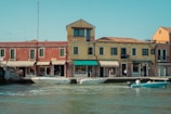 A picturesque scene of a waterfront with colorful Italian-style buildings and several people leisurely walking along the sidewalk. Two boats with passengers are navigating the calm waters in front of the buildings that feature rustic facades and vibrant awnings. The sky is clear, indicating a pleasant day.