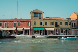 A picturesque scene of a waterfront with colorful Italian-style buildings and several people leisurely walking along the sidewalk. Two boats with passengers are navigating the calm waters in front of the buildings that feature rustic facades and vibrant awnings. The sky is clear, indicating a pleasant day.