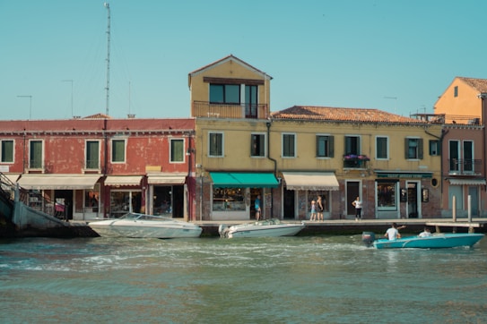 A picturesque scene of a waterfront with colorful Italian-style buildings and several people leisurely walking along the sidewalk. Two boats with passengers are navigating the calm waters in front of the buildings that feature rustic facades and vibrant awnings. The sky is clear, indicating a pleasant day.