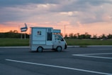 A small street bar truck labeled 'Frizzy & Spritzy' is driving along an open road during sunset, with a grassy field and trees in the background and a partly cloudy sky.