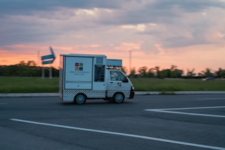 A small street bar truck labeled 'Frizzy & Spritzy' is driving along an open road during sunset, with a grassy field and trees in the background and a partly cloudy sky.