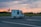 A small street bar truck labeled 'Frizzy & Spritzy' is driving along an open road during sunset, with a grassy field and trees in the background and a partly cloudy sky.
