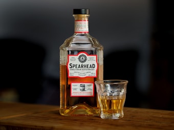 A clear bottle of Spearhead Single Grain Scotch Whisky stands on a wooden surface with a filled glass beside it. The bottle features a label with red and white colors, displaying the brand name and product details. The background is blurred, emphasizing the bottle and glass as the main focus.