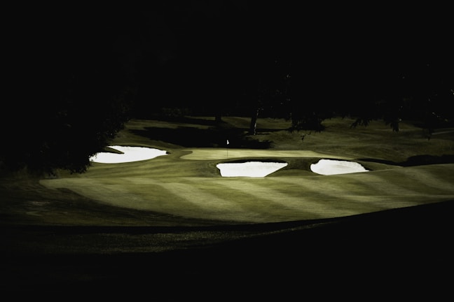 Dark, moody golf course scene with sand traps and a fairway under dim light conditions, creating strong shadows and contrasts.