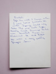 A handwritten note in blue ink on a white piece of paper. The handwriting is cursive and slightly slanted to the right. The background is a plain, soft pink surface.
