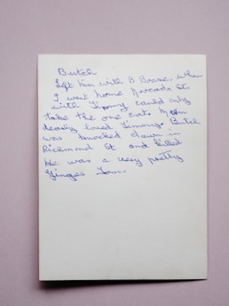 A handwritten note in blue ink on a white piece of paper. The handwriting is cursive and slightly slanted to the right. The background is a plain, soft pink surface.