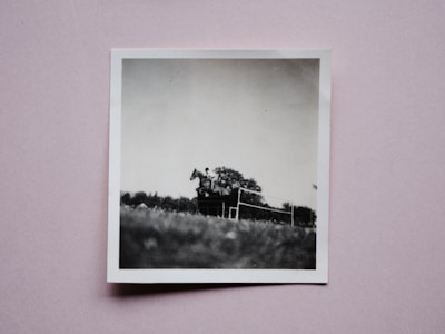 A black and white photograph featuring a person riding a horse as it jumps over a fence. The scene is captured in an outdoor setting with trees in the background and a focus on the equestrian activity. The photograph is a physical print laid on a pink background.