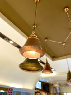 Hanging bell-shaped pendant lights with a metallic finish, suspended from the ceiling of a modern interior space. The lights are on, casting a warm glow. The background features blurred elements of a contemporary setting with display shelves and screens.