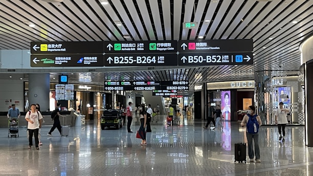 An airport terminal hallway with several people walking, some pulling luggage. Overhead are illuminated directional signs with text in multiple languages indicating directions to gates, baggage claim, and other airport facilities. Luxury brand storefronts such as Gucci are visible on the sides.