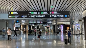 An airport terminal hallway with several people walking, some pulling luggage. Overhead are illuminated directional signs with text in multiple languages indicating directions to gates, baggage claim, and other airport facilities. Luxury brand storefronts such as Gucci are visible on the sides.