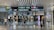 An airport terminal hallway with several people walking, some pulling luggage. Overhead are illuminated directional signs with text in multiple languages indicating directions to gates, baggage claim, and other airport facilities. Luxury brand storefronts such as Gucci are visible on the sides.