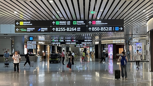 An airport terminal hallway with several people walking, some pulling luggage. Overhead are illuminated directional signs with text in multiple languages indicating directions to gates, baggage claim, and other airport facilities. Luxury brand storefronts such as Gucci are visible on the sides.