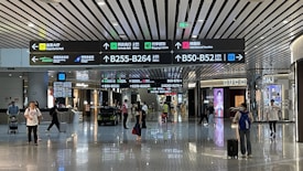 An airport terminal hallway with several people walking, some pulling luggage. Overhead are illuminated directional signs with text in multiple languages indicating directions to gates, baggage claim, and other airport facilities. Luxury brand storefronts such as Gucci are visible on the sides.
