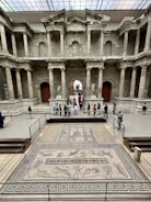 A large ancient stone structure featuring intricate architectural details such as columns, arches, and carvings. In the foreground, there is an ornate mosaic floor with detailed patterns and figures. Several people are standing in the area, observing the historical site.