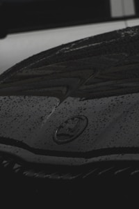 A close-up view of a car hood with water droplets scattered across its surface. The logo of the car is visible in the center, surrounded by the dark, glossy finish of the vehicle.