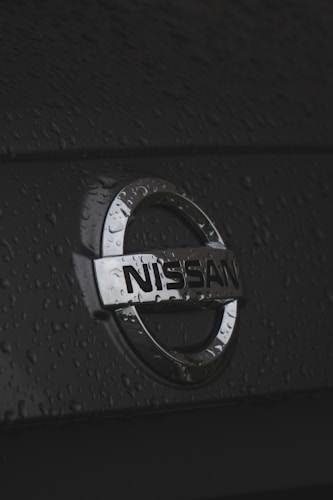 A close-up view of the Nissan logo on the back of a car, with water droplets scattered across the surface. The metal emblem appears shiny against the dark background, with raindrops adding a textured effect.
