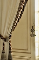 A decorative cream-colored curtain draped elegantly with a braided tieback featuring intricate gold and brown details. The curtain is hanging beside a vintage-style wooden door with ornate hardware, allowing soft natural light to filter through.