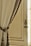 A decorative cream-colored curtain draped elegantly with a braided tieback featuring intricate gold and brown details. The curtain is hanging beside a vintage-style wooden door with ornate hardware, allowing soft natural light to filter through.