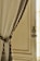 A decorative cream-colored curtain draped elegantly with a braided tieback featuring intricate gold and brown details. The curtain is hanging beside a vintage-style wooden door with ornate hardware, allowing soft natural light to filter through.