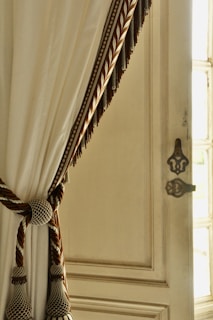 A decorative cream-colored curtain draped elegantly with a braided tieback featuring intricate gold and brown details. The curtain is hanging beside a vintage-style wooden door with ornate hardware, allowing soft natural light to filter through.