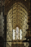 Intricate wrought iron scrollwork creates an ornate pattern. The background is softly lit, featuring stained glass that diffuses warm light, enhancing the elegance and complexity of the metalwork.