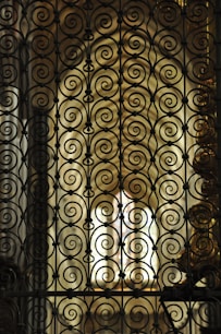 Intricate wrought iron scrollwork creates an ornate pattern. The background is softly lit, featuring stained glass that diffuses warm light, enhancing the elegance and complexity of the metalwork.