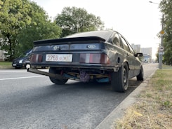 A black Ford car is parked on the side of a road. The rear view highlights the license plate and the exhaust system, which protrudes noticeably. The car appears to have a worn and modified look, with visible customizations under the bumper. Nearby, there are trees and another vehicle parked down the street. Buildings and a clear sky can be seen in the background.