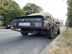 A black Ford car is parked on the side of a road. The rear view highlights the license plate and the exhaust system, which protrudes noticeably. The car appears to have a worn and modified look, with visible customizations under the bumper. Nearby, there are trees and another vehicle parked down the street. Buildings and a clear sky can be seen in the background.