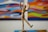 A wooden mannequin is posed in a dynamic and playful stance with one leg lifted and arms raised. It stands in front of a colorful abstract painting that features swirling patterns and vibrant colors.