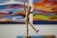 A wooden mannequin is posed in a dynamic and playful stance with one leg lifted and arms raised. It stands in front of a colorful abstract painting that features swirling patterns and vibrant colors.