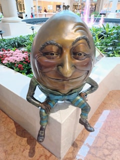 A whimsical statue of an egg-headed character with a humorous expression is sitting on a ledge. The character is dressed in colorful, striped clothing and has exaggerated facial features, including a large nose and sly smile. Surrounding the figure are lush green plants and bright pink flowers. In the distance, a water feature is visible, and the interior of a shopping mall can be seen with people and stores.