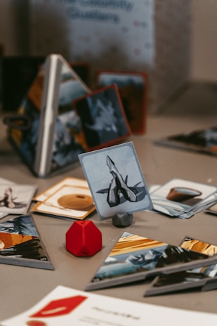A tabletop game setup with various illustrated cards displayed on a surface. A red, geometric game piece is positioned next to the cards, adding a pop of color. The cards have images and symbols, suggesting a fantasy or strategy theme. The background shows a partially obscured game box with text.