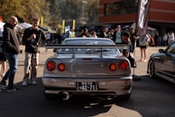 A parking lot gathering featuring a silver sports car with a large rear spoiler. People are gathered around, socializing in a casual setting. Bright sunlight and colorful tents indicate a lively car meet or event.