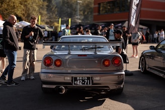 A parking lot gathering featuring a silver sports car with a large rear spoiler. People are gathered around, socializing in a casual setting. Bright sunlight and colorful tents indicate a lively car meet or event.
