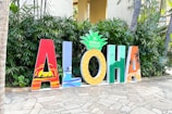 Large colorful letters spelling out 'ALOHA' are displayed on a stone pathway. Each letter features a distinct design, incorporating tropical and beach-themed elements such as palm trees and surfers. The letter 'O' is styled as a pineapple with the Hilton Hawaiian Village logo on top. Green foliage serves as a backdrop.