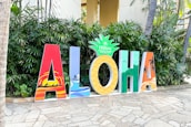 Large colorful letters spelling out 'ALOHA' are displayed on a stone pathway. Each letter features a distinct design, incorporating tropical and beach-themed elements such as palm trees and surfers. The letter 'O' is styled as a pineapple with the Hilton Hawaiian Village logo on top. Green foliage serves as a backdrop.