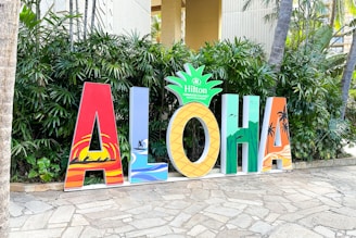 Large colorful letters spelling out 'ALOHA' are displayed on a stone pathway. Each letter features a distinct design, incorporating tropical and beach-themed elements such as palm trees and surfers. The letter 'O' is styled as a pineapple with the Hilton Hawaiian Village logo on top. Green foliage serves as a backdrop.