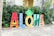 Large colorful letters spelling out 'ALOHA' are displayed on a stone pathway. Each letter features a distinct design, incorporating tropical and beach-themed elements such as palm trees and surfers. The letter 'O' is styled as a pineapple with the Hilton Hawaiian Village logo on top. Green foliage serves as a backdrop.