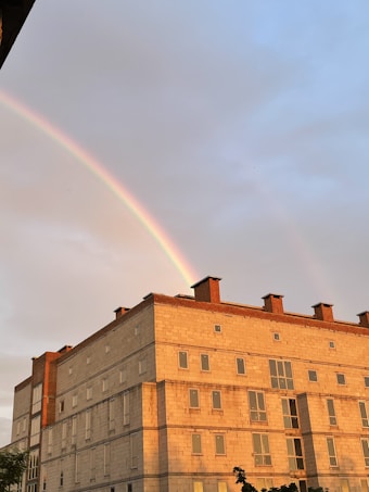 A rainbow gracefully arcs across the sky above a large brick building with multiple windows. The building has a modern architectural style with a flat roof and is partially illuminated by sunlight. The sky transitions from cloudy to clear, offering a mix of warm and cool tones.