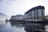 A large, elegant hotel building with multiple floors and balconies is situated alongside a calm body of water, possibly a lake. The scene is set in a mountainous area with misty hills in the background and a clear sky above. The architecture is traditional with a combination of white and beige colors, and there are small decorative plants on the balconies. The water reflects the building and surrounding landscape, adding to the tranquil atmosphere.